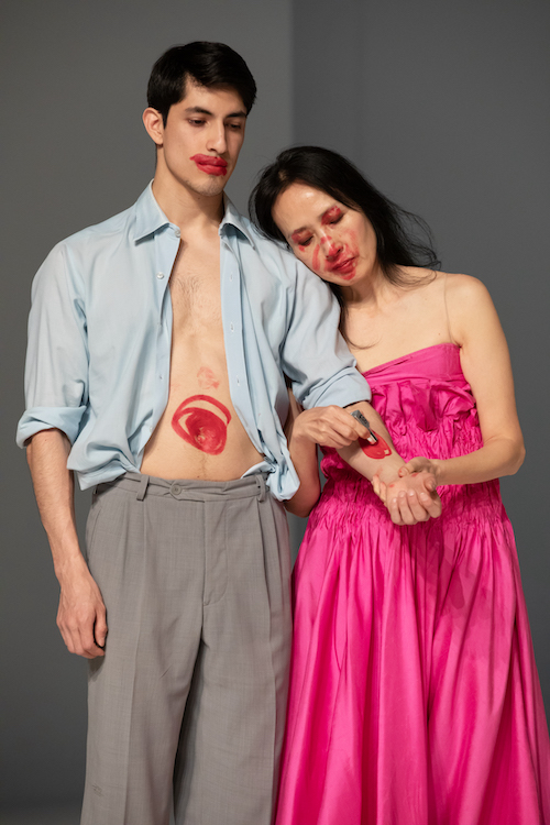 close up of a couple, man and woman head and torso shot. She leans on his shoulder and draws a lipstick circle on his forearm. He has lipstick messily applied on his lips and on his belly, she has a circle of lipstick on her nose and mouth. It looks like war paint. 